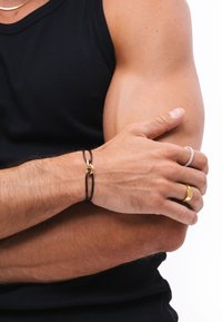 Black leather bracelet with gold hardware, worn on a hand; additional rings visible in silver and gold tones. Background is white.