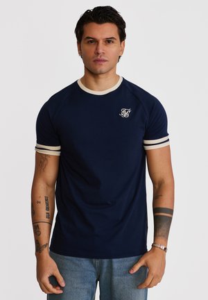 Uomo con capelli scuri che indossa una maglietta blu navy con rifiniture bianche e logo, jeans azzurri e tatuaggi visibili su entrambe le braccia.