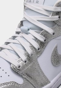 Nærbilde av hvit og metallisk sølv joggesko med hvite lisser og strukturert Nike-logo på siden.