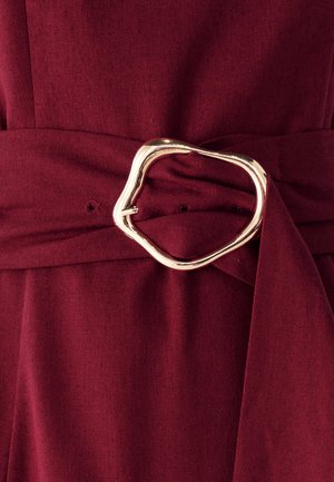 Burgundi värvi kangas, millel on sile tekstuur, omab lai vöö, mis on seotud vöökoha ümber ning millel on ebaühtlane kuldset tooni metallist pandla aktsent.