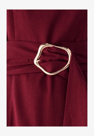 Bordeauxfarbener Stoff mit glatter Textur verfügt über einen breiten Gürtel, der um die Taille gebunden ist und von einer unregelmäßigen goldfarbigen Metallschnalle akzentuiert wird.