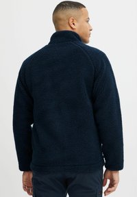 Marineblauwe fleece jack met een zachte, gestructureerde afwerking, hoge kraag en lange mouwen, gezien vanuit de achterkant. Geen zichtbare branding of accenten.
