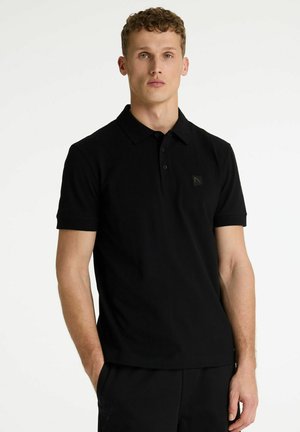 Joven con camisa polo negra de manga corta y un pequeño logo en el pecho, de pie frente a un fondo blanco liso.