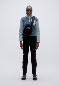 Φλις πουλόβερ σε μπλε και γκρι, μαύρο σακίδιο περασμένο στον ώμο, μαύρο παντελόνι και ανθεκτικά μαύρα παπούτσια· αθλητικά γυαλιά ηλίου ολοκληρώνουν το ντύσιμο.