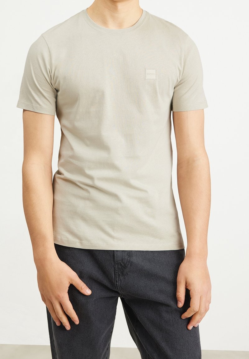 Homme portant un t-shirt uni beige clair à manches courtes et un pantalon gris foncé, debout, les bras détendus le long du corps.