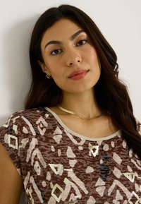 Camiseta de manga corta marrón con patrones geométricos abstractos en blanco y metálico. Tiene un escote redondeado y está fabricada con una tela suave y ligera.