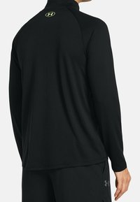 Schwarzes langärmliges Performance-Shirt mit hohem Kragen. Mit einem leuchtend gelben Logo auf dem Rücken und einer glatten, feuchtigkeitsableitenden Stoffstruktur.