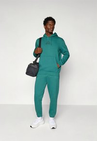 Tirkīza krāsas sporta džemperis ar kengura kabatu un logotipu, komplektā ar atbilstošām treniņbiksēm. Valkāts ar baltām sporta kurpēm un melnu pastnieka somu.