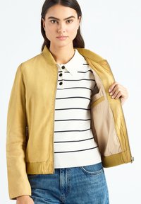 Blouson aviateur en cuir jaune avec col et poignets côtelés, fermeture éclair à l'avant et doublure intérieure lisse. Polo rayé blanc et bleu marine en dessous.