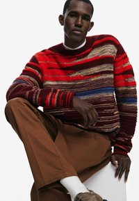 Rot gemusterter Pullover mit horizontalen Streifen in verschiedenen Farben, kombiniert mit braunen Hosen. Die Stricktextur und der Rundhalsausschnitt sind deutlich sichtbar.