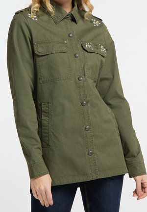 Kvinde iført en olivengrøn button-up jakke med blomsterbroderi på skuldrene og frontlommerne, kombineret med mørke jeans.