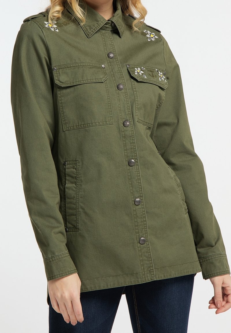 Femme portant une veste boutonnée vert olive avec une broderie florale sur les épaules et les poches avant, assortie à un jean foncé.