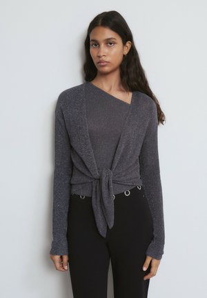 Graue, funkelnde Strickjacke mit langen Ärmeln, vorne zum Binden. Getragen über einem passenden, gerippten, figurbetonten Top. Schwarze Hose mit Ösenakzenten.