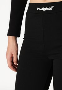 Leggings noirs avec une taille haute portant le logo "lowlights" en blanc. Le tissu semble extensible et lisse avec des coutures minimales.