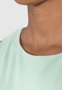 Top verde menta con escote redondo, hecho de una tela suave, mostrando un primer plano del área del hombro y el cuello. Sin herrajes ni patrones visibles.