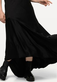 Robe maxi en satin noir avec ourlet asymétrique, présentant une silhouette fluide. Associée à des bottines noires robustes. Texture lisse.