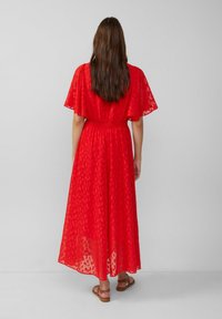 Vrouw met lang bruin haar, gekleed in een rode gemêleerde midi-jurk en brown sandalen, staand tegen een effen lichtgrijze achtergrond.