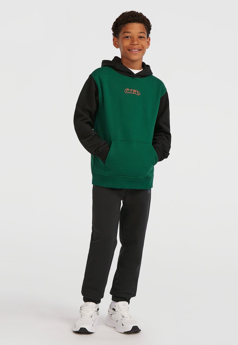 Green and black hooded sweatshirt with a front pocket and graphic logo, paired with black tapered sweatpants and white athletic shoes.