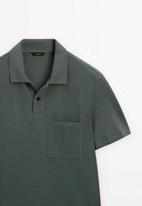 Polo vert foncé à manches courtes avec col, un seul bouton noir et une poche carrée sur la poitrine sur fond blanc.