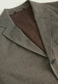 Nahaufnahme eines maßgeschneiderten Blazers mit einem strukturierten grauen Muster und brauner Fütterung, die den Kragen und die Knopfdetails zeigt.