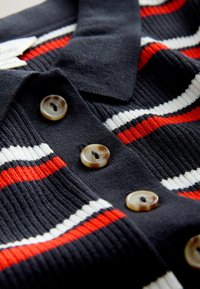 Polo en maille bleu marine avec des rayures rouges et blanches, doté de quatre boutons ronds marbrés et d'un col classique. Texture douce.