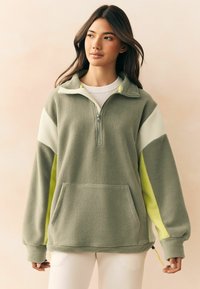 Fleece tipo megztinis ramiai žalia spalva su baltomis ir neoninėmis geltonomis detalėmis. Turi pusiau užtrauktuką, aukštą apykaklę ir priekinę kišenę su švelnia tekstūra.