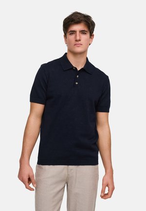 Navy poloshirt met korte mouwen, zachte textuur, knoopsluiting met drie knopen en geribbelde zoom. Draagt zich met lichtgekleurde broek.