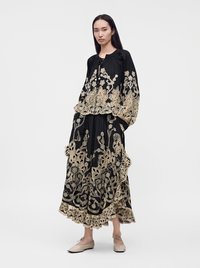 Femme portant un haut à manches longues noir et une jupe avec une broderie florale dorée, debout devant un fond clair uni.