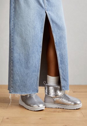Jambes portant une jupe en denim bleu clair avec une fente haute, des chaussettes blanches et des bottines argentées brillantes à sequins sur un sol en bois.