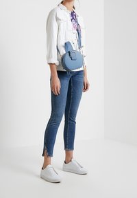 Blauwe ronde crossbody tas met een bovenhandvat, zachte textuur, gouden hardware en een verstelbare schouderband; gedragen met een witte jas en blauwe spijkerbroek.