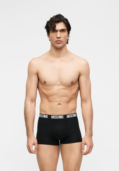 Άνδρας χωρίς πουκάμισο που φοράει μαύρα boxer briefs Moschino στέκεται μπροστά σε απλό γκρι φόντο, κοιτάζοντας ευθεία με ουδέτερη έκφραση.