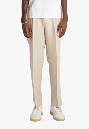 Uomo che indossa pantaloni beige su misura, sneakers bianchi e una camicia bianca, in piedi con le mani rilassate lungo i fianchi su uno sfondo bianco.