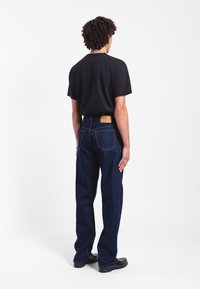 Dark blue straight-leg denim jeans with front and back pockets, featuring a tan leather patch at the waist and subtle yellow stitching.