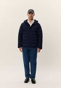 Άνδρας που φοράει μαύρο καπέλο, μπλε σκούρο μπουφάν φουσκωτό, μπεζ πουλόβερ με φερμουάρ, μπλε τζιν και μαύρα επίσημα παπούτσια, στέκεται μπροστά σε ένα φωτεινό φόντο.