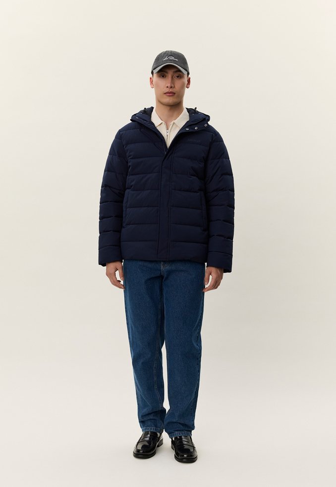 Άνδρας που φοράει μαύρο καπέλο, μπλε σκούρο μπουφάν φουσκωτό, μπεζ πουλόβερ με φερμουάρ, μπλε τζιν και μαύρα επίσημα παπούτσια, στέκεται μπροστά σε ένα φωτεινό φόντο.