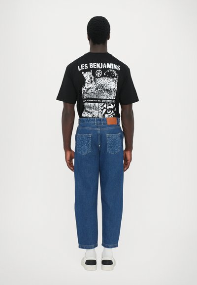 Μαύρο γραφικό μπλουζάκι με λευκή εκτύπωση, μπλε τζιν παντελόνι με υφή στις πίσω τσέπες και λευκά αθλητικά παπούτσια με μαύρες λεπτομέρειες.