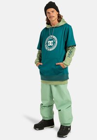 Turkusowa bluza z dużym logo, założona na zieloną bluzkę z długim rękawem. Uzupełniona o jasne zielone spodnie i czarne śniegowce. Na głowie czarna czapka.