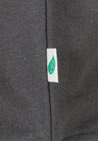 Sötétszürke szövet sima textúrával, egy fehér címkével, zöld levél logóval, a ruhadarab szélén elhelyezve.