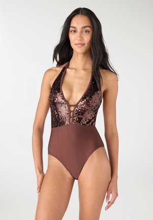 Szczupła kobieta w brązowym stroju kąpielowym z dekoltem halter, ozdobionym cekinami na górze oraz głębokim wycięciem w kształcie litery V, stojąca na jednolitym tle.