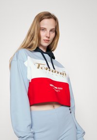 Strihnutá mikina s kapucňou vo svetlomodrej, bielej a červenej farbe. Obsahuje zlatý logo text a nápis "NEW YORK CITY" vytlačený na bielo. Mäkká textúra, neformálny dizajn.
