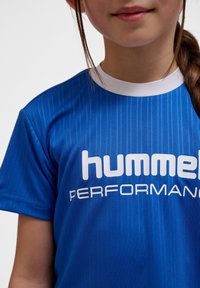 Blå sporttröja med vertikala ränder, vit krage och "hummel PERFORMANCE" logga i vitt. Tyget verkar slätt och lätt.