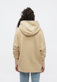 Μπεζ φλις φούτερ με μεγάλο, στρογγυλεμένο κουκούλι, χαμηλωμένους ώμους και απαλή υφή, συνδυασμένο με ανοιχτόχρωμο μπλε τζιν.