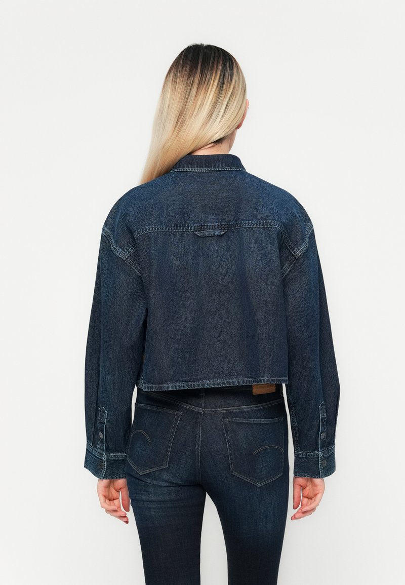 Afklippet denimjakke i mørkeblå, med lange ærmer, knaplukning ved håndleddene og lige kant. Ensfarvet ryg med subtile stikningsdetaljer.