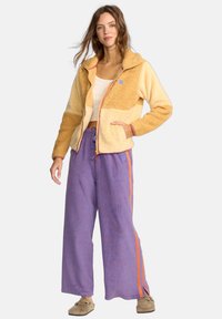 Fleece jacket in yellow and beige with a zip, paired with purple striped wide-leg pants featuring orange accents. Soft, textured fabric.