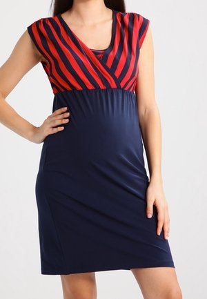 Frau trägt ein ärmelloses Umstandskleid mit roten und marineblauen diagonalen Streifen im oberen Bereich und einem einfarbigen marineblauen Rock, steht mit der Hand in der Hüfte.