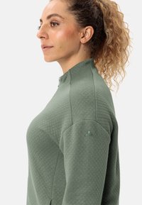 Pullover imbottito verde con colletto alto e zip. Presenta un ricamo del logo sottile sulla spalla. Tessuto morbido e testurizzato.
