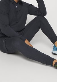 Haut de sport gris foncé avec une poche kangourou, un jogging assorti et des chaussures de sport bleues. Le matériau semble doux avec une texture lisse.