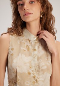 Blusa sin mangas en beige claro con estampado floral, presenta un cuello y un cierre de botones. La tela tiene una apariencia suave y texturizada.