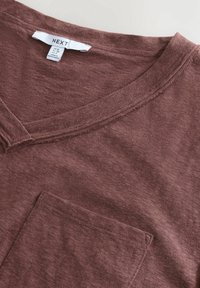 Bordowy t-shirt z dekoltem w serek, wykonany z miękkiego materiału, z kieszonką z przodu i surowym wykończeniem dekoltu. Metka widoczna wewnątrz kołnierza.