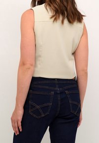 Žena v bezramenném béžovém topu a tmavě modrých džínách, viděná z pohledu zezadu, stojící na pozadí s jednolitým světlým pozadím.